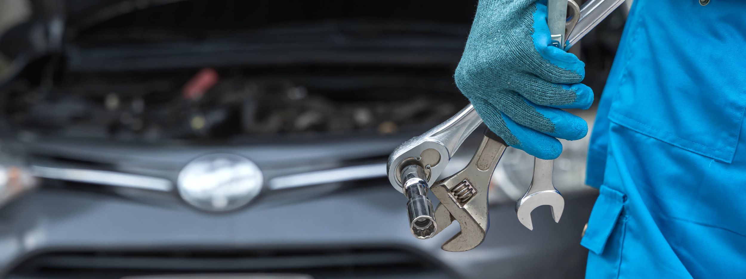 Eine Hand eines Mechanikers mit Werkzeugen vor einer geöffneten Motorhaube.